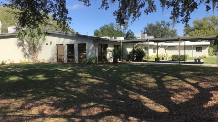 Exterior view of cream-colored residential building with covered pavilion, manicured lawn, palm trees, and natural shade