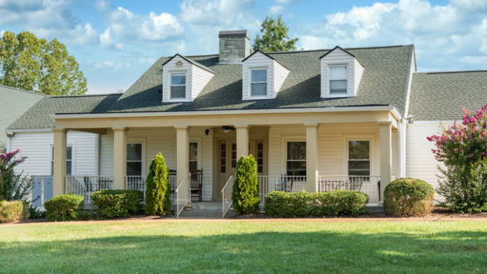 Exterior of yellow house with white covered porch, dormer windows, shutters, landscaping, and manicured lawn