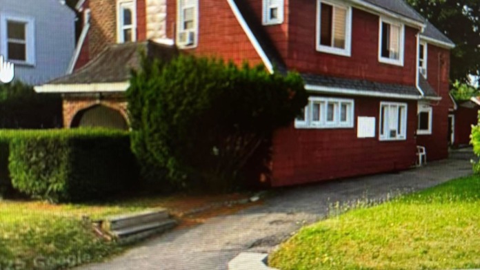 Exterior of red brick residential house with peaked roof, chimney, manicured landscaping, driveway, and stone retaining wall.