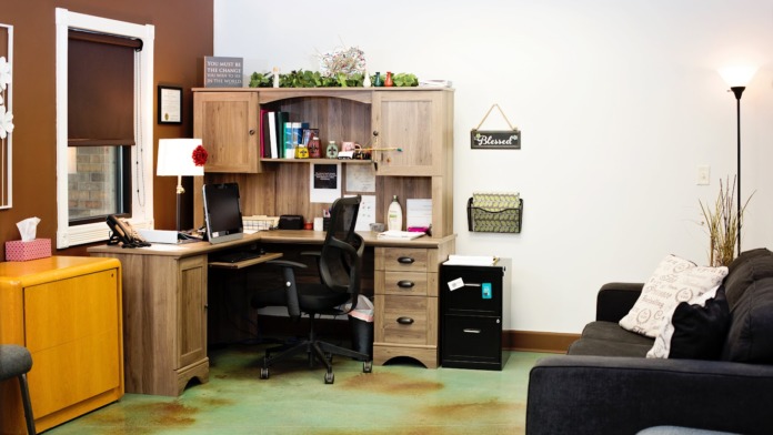 Interior bedroom-study combination with wooden desk, office chair, filing cabinets, sofa seating, and storage shelving with decorative plants