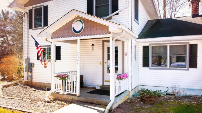 Exterior view of white residential-style building entrance with covered porch, decorative shutters, American flag, and flowering plants