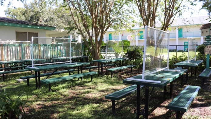 Outdoor picnic area with green benches, white canopy structure, and tall trees providing shade