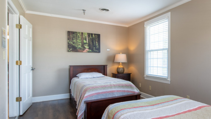 Interior bedroom with two beds, wood nightstand, lamp, window with blinds, and white closet doors
