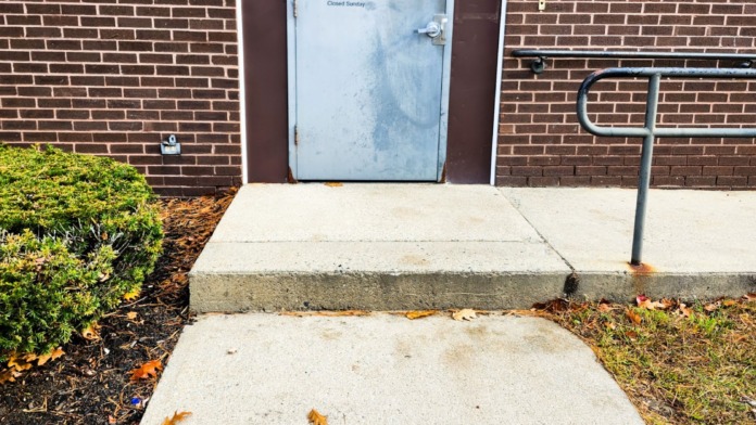 Exterior brick wall showing metal-framed entrance door with facility hours and contact information signage, concrete walkway, hand railing