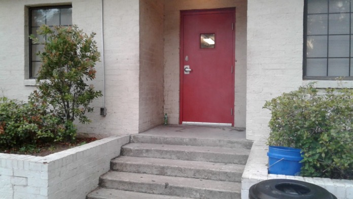 Exterior entryway with red door, concrete steps, white brick, plantings, and receptacle