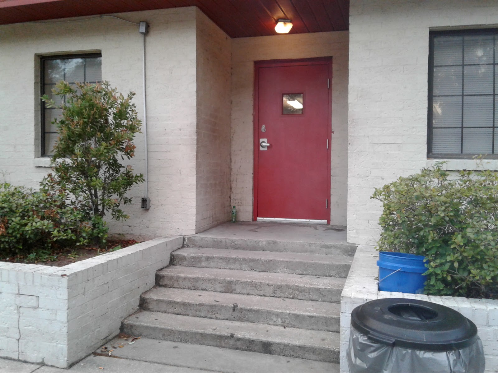 Exterior entryway with red door, concrete steps, white brick, plantings, and receptacle