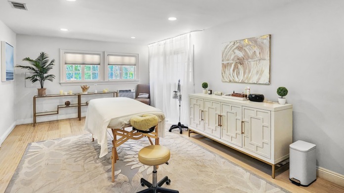 Interior spa treatment room with massage table, cream furnishings, fireplace, windows, and ocean-facing balcony