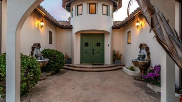 Exterior arched entryway with white stucco walls, decorative tile roof, green wooden doors, stone courtyard and landscaping
