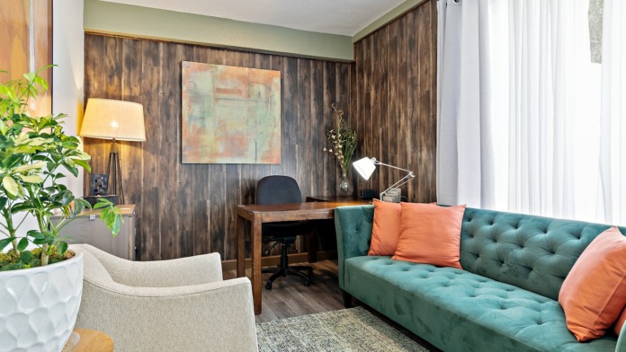 Interior office space with wooden desk, ergonomic chair, computer monitor, mirror, plants, and bright windows overlooking grounds