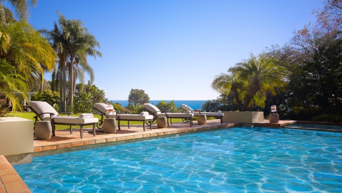 Outdoor swimming pool with lounge chairs, ocean view, and palm tree landscaping in background