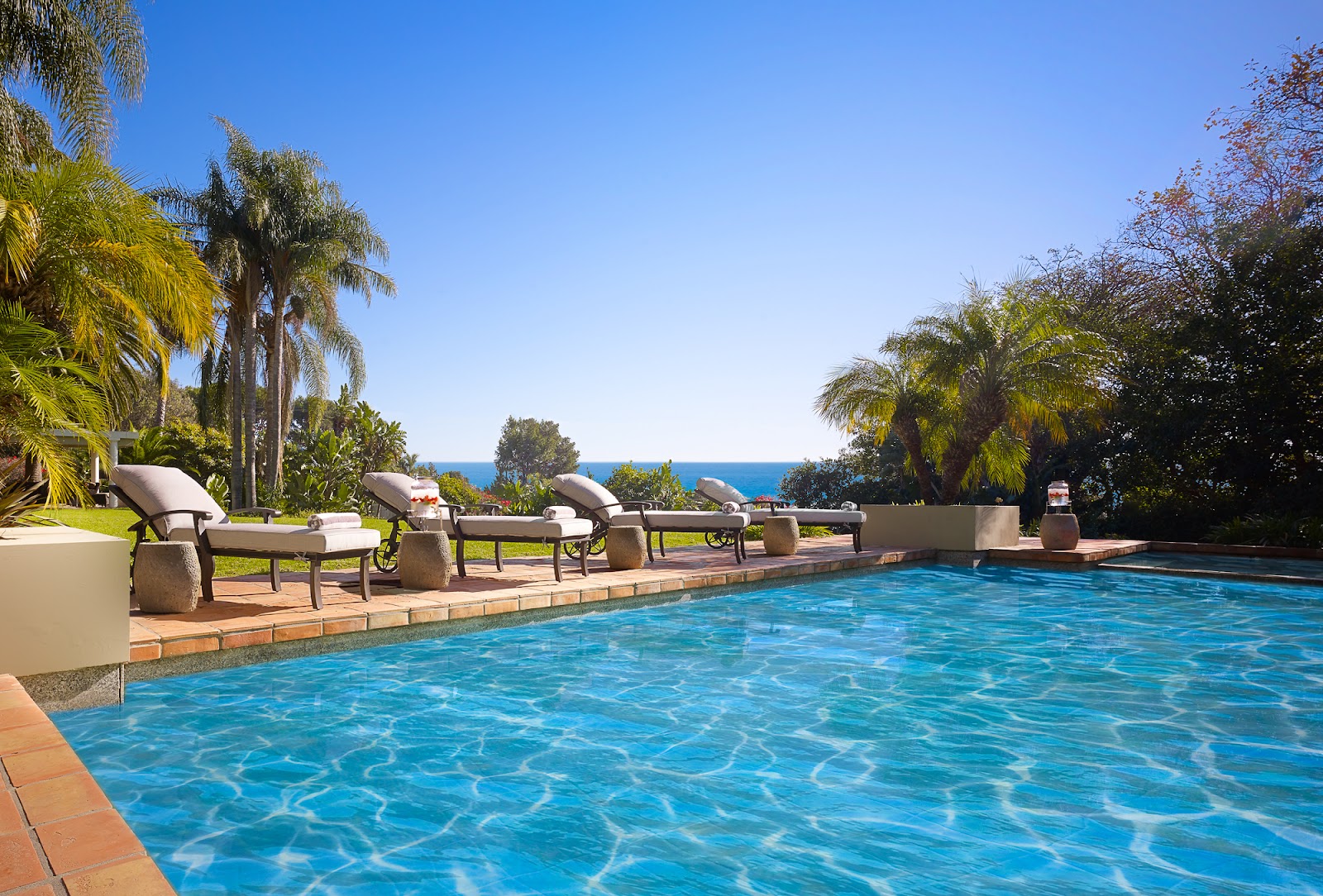 Outdoor swimming pool with lounge chairs, ocean view, and palm tree landscaping in background