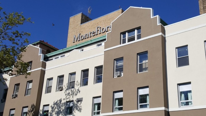 Exterior tall multi-story tan and white hospital building with Montefiore roofline signage and street-level storefront entrance