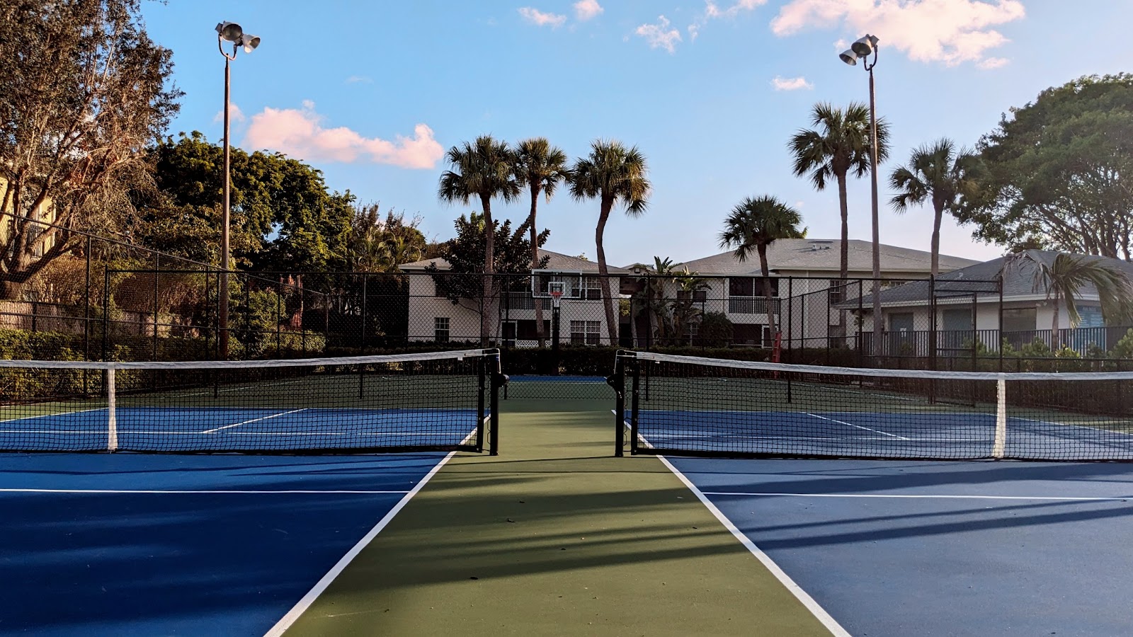 Outdoor tennis courts with blue and green playing surfaces, white nets, palm trees, and facility buildings in background
