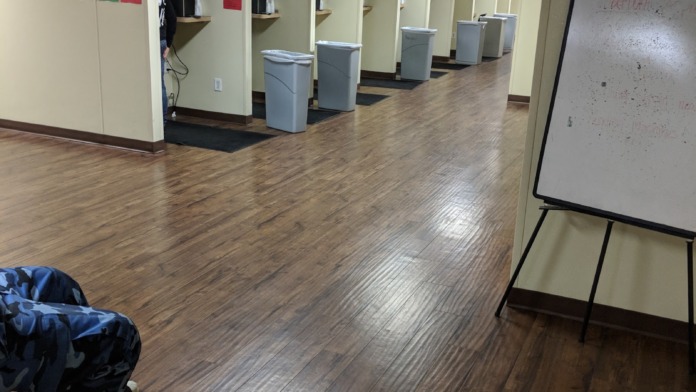 Interior hallway with multiple partitioned assessment booths, gray bins, wooden flooring, and information monitors
