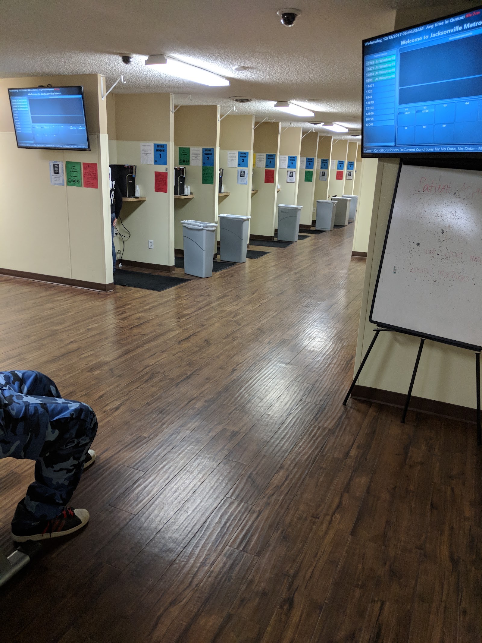 Interior hallway with multiple partitioned assessment booths, gray bins, wooden flooring, and information monitors