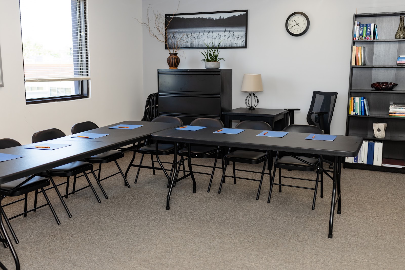 Interior meeting space with long conference table, multiple office chairs, bookshelf, clock, and window