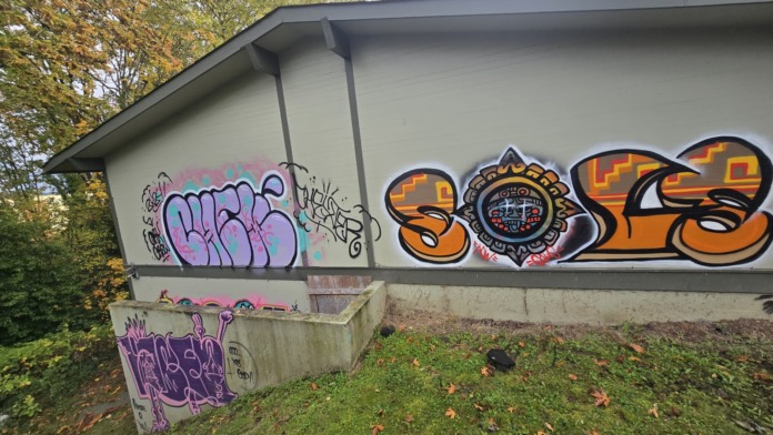 Exterior view of facility building with graffiti on facade, courtyard seating area, and landscaped grounds surrounded by mature trees