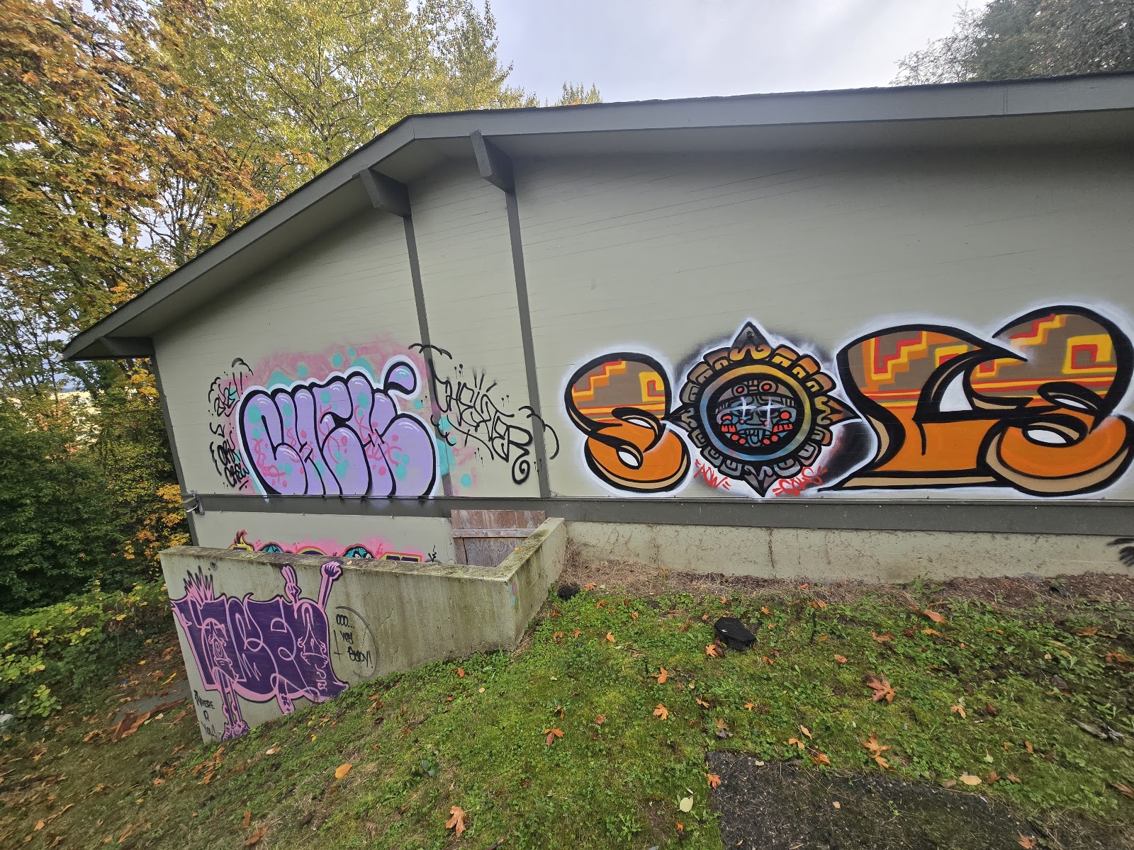 Exterior view of facility building with graffiti on facade, courtyard seating area, and landscaped grounds surrounded by mature trees
