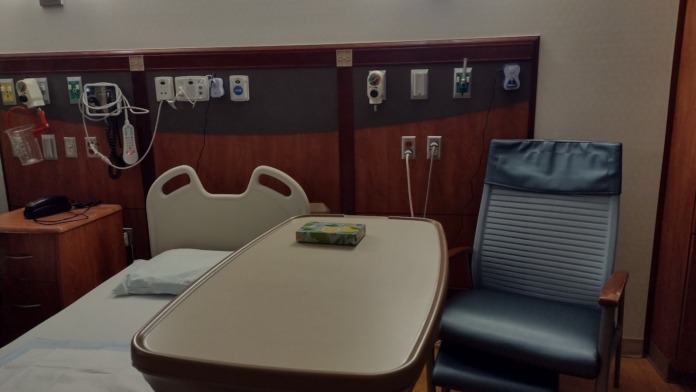 Interior medical treatment room with hospital bed, visitor chair, wall-mounted medical equipment and outlets, clock, and window with natural light