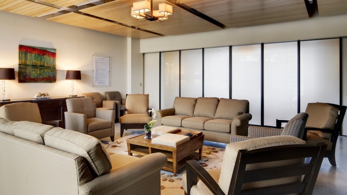Interior lounge with tan upholstered furniture, wooden coffee table, modern lighting, windows with sheer curtains and framed wall art.