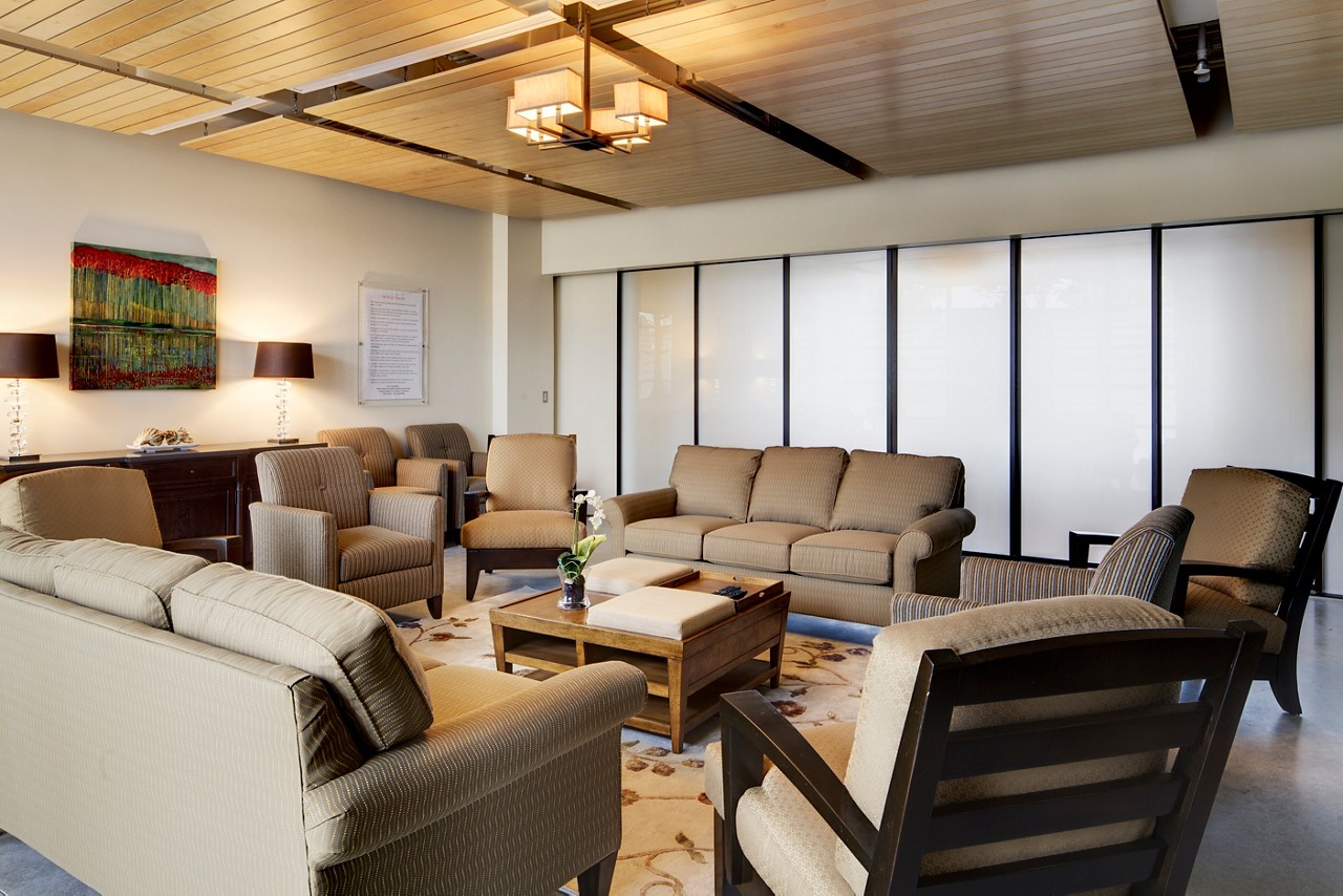 Interior lounge with tan upholstered furniture, wooden coffee table, modern lighting, windows with sheer curtains and framed wall art.