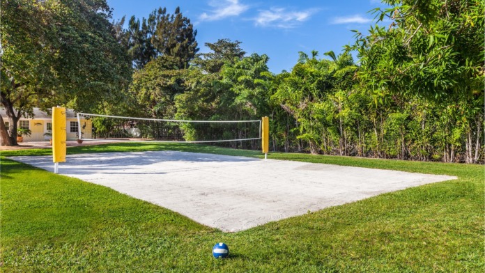 Outdoor recreational volleyball court with sand surface, yellow net supports, green lawn, and dense tree line backdrop