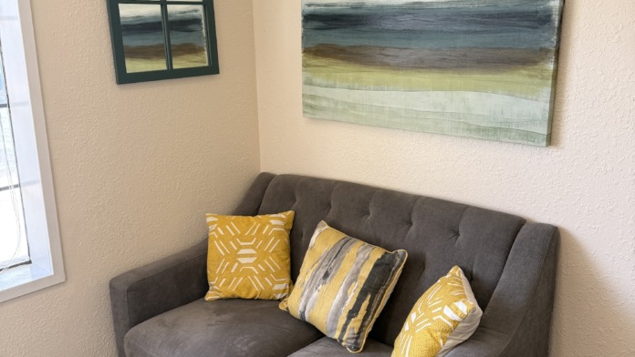 Interior seating nook with gray upholstered loveseat, decorative cushions, landscape artwork, and window frame decoration.