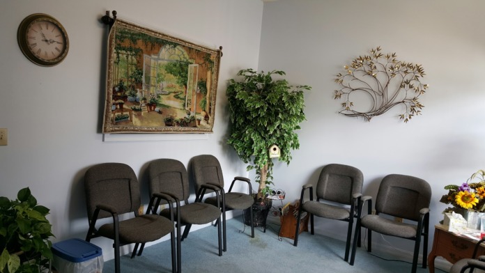 Interior waiting room with gray upholstered chairs arranged around walls, decorated with wall art, plants, and flowers
