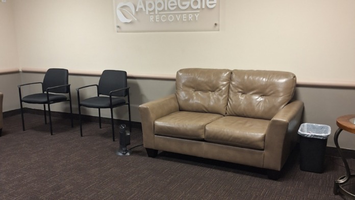 Interior waiting area with AppleGate Recovery wall sign, tan sofa, black seating, and brown carpet flooring.