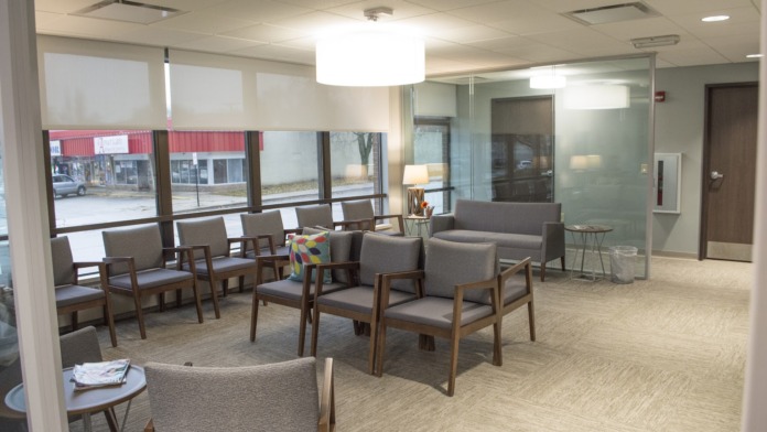 Interior waiting area with multiple gray chairs, large windows, glass partitions, and recessed ceiling lights.