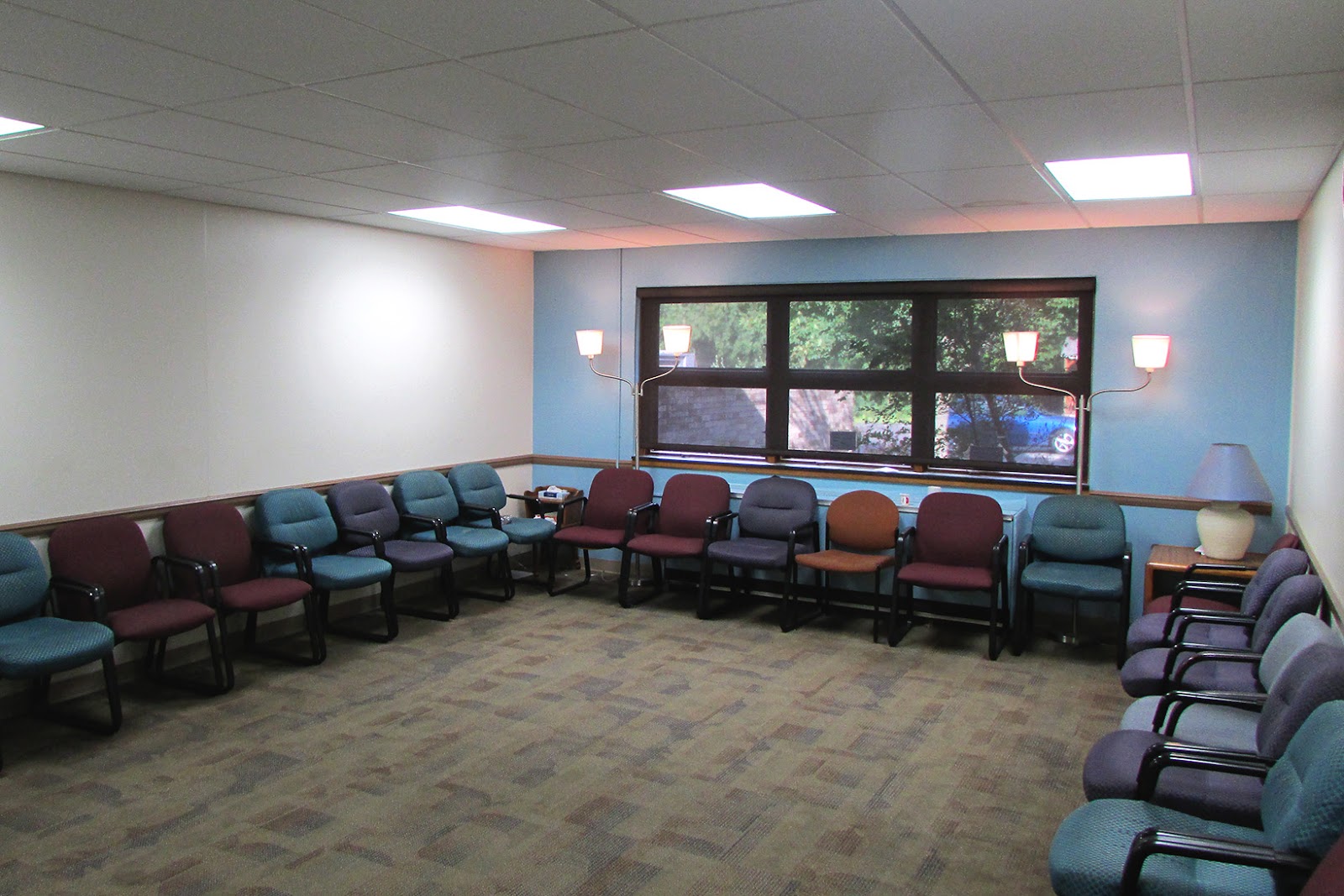 Interior waiting area with arranged upholstered chairs in burgundy and teal, recessed lighting, and large windows.