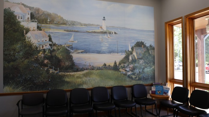 Interior waiting area with row of black chairs facing large coastal landscape mural with sailboats and lighthouse scene