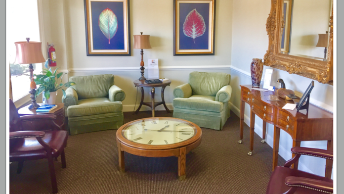 Interior waiting room with green armchairs, round center table with clock, framed leaf artwork, and wood furnishings