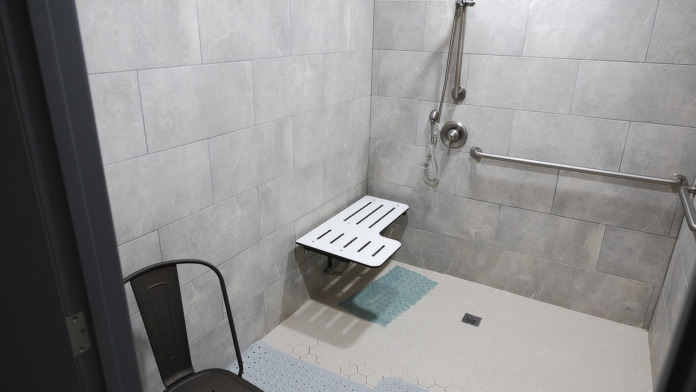 Interior walk-in shower with handrails, mat, accessible controls, beige tile, and safety features for patients