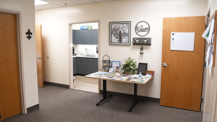 Interior entry with wooden doors, information table, materials display, and kitchen visible through glass.