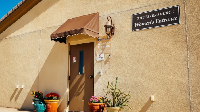 Exterior women's program entrance with brown door, canopy, flowering planters in terra cotta and ceramic pots, tan stucco building