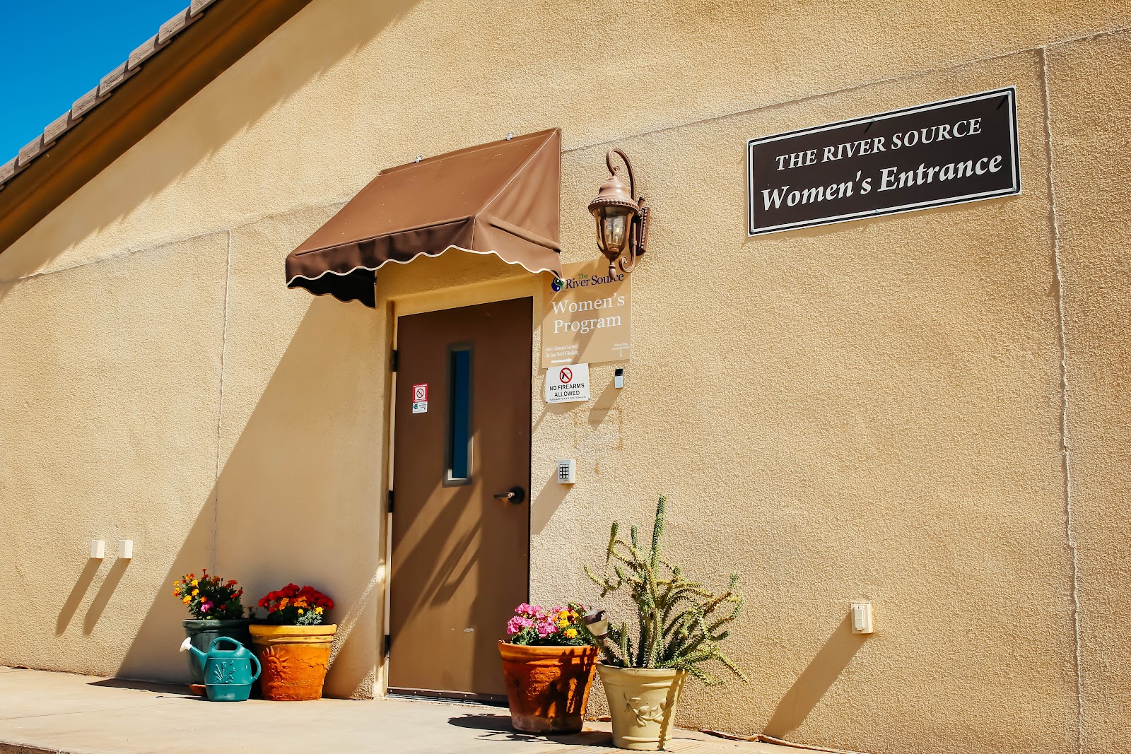Exterior women's program entrance with brown door, canopy, flowering planters in terra cotta and ceramic pots, tan stucco building