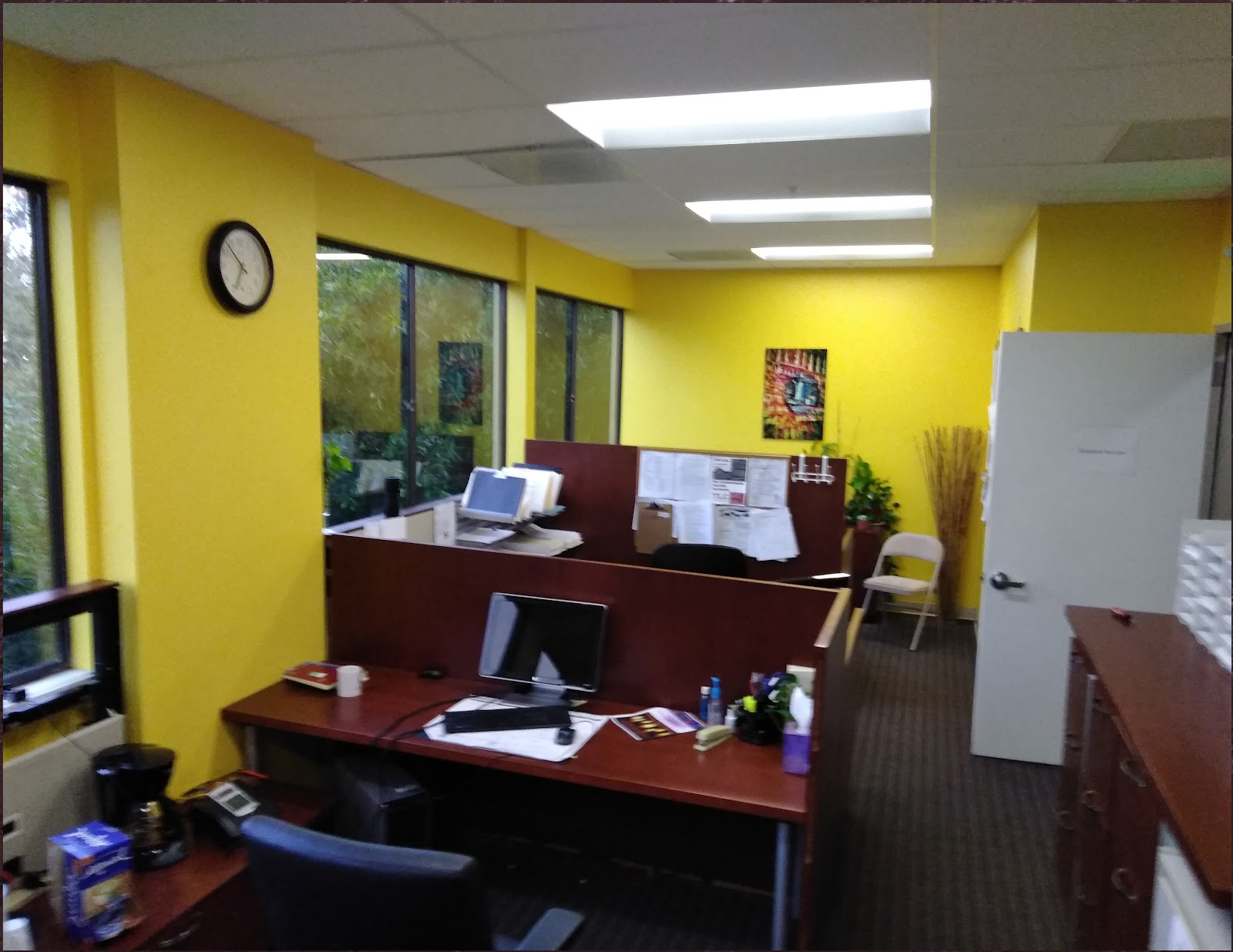 Interior office with yellow walls, wooden desks, computers, and natural window lighting.
