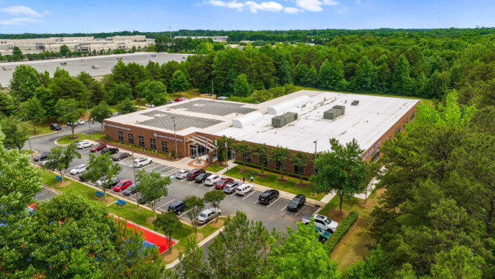 Aerial view of brick facility building with white roof, parking areas, mature landscaping, and surrounding commercial district