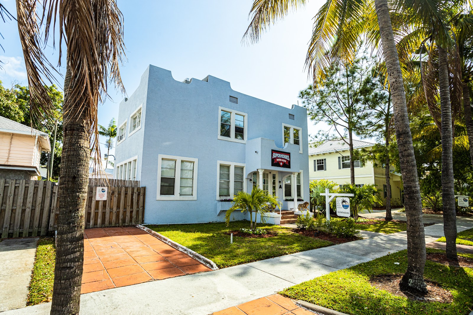 Exterior front facade of two-story blue building with white trim, brown brick entrance steps, palm trees, and landscaping.