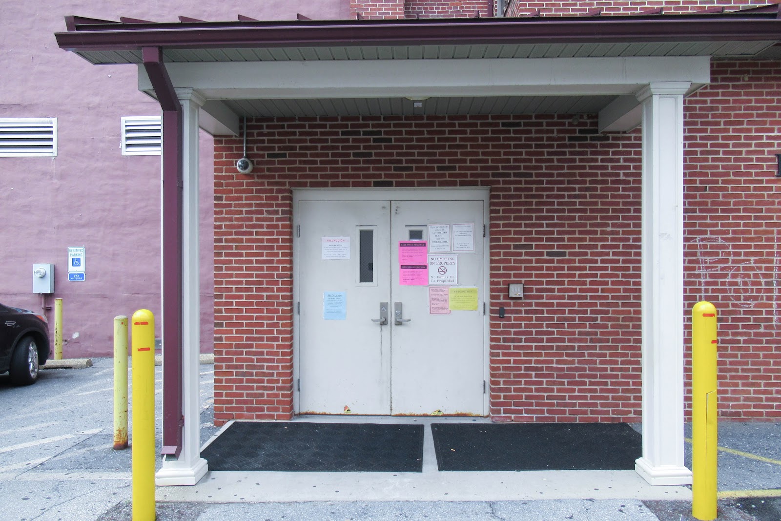 Exterior entrance with white double doors, yellow protective bollards, covered overhang, and brick construction