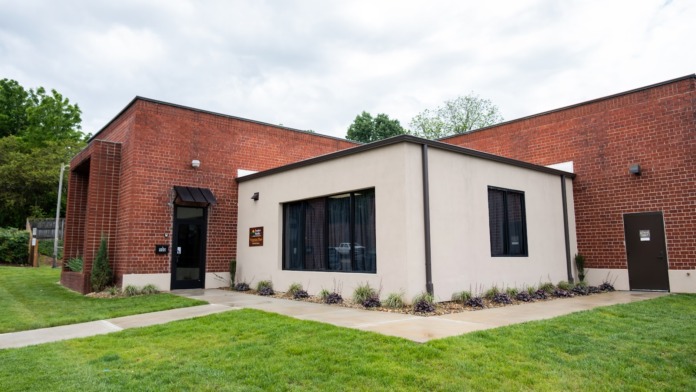 Exterior of brick building with modern white addition, manicured lawn, concrete pathways, and multiple entry doors.