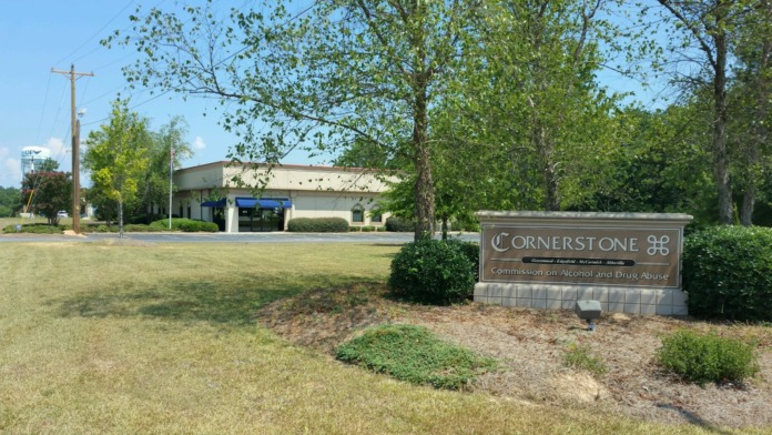 Exterior view showing tan and blue commercial structure, mature trees, utility infrastructure, and manicured lawn area.