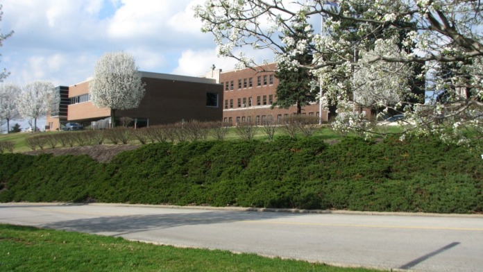 Exterior view of multi-building facility complex with brick structures, manicured hedges, and seasonal flowering trees in spring.
