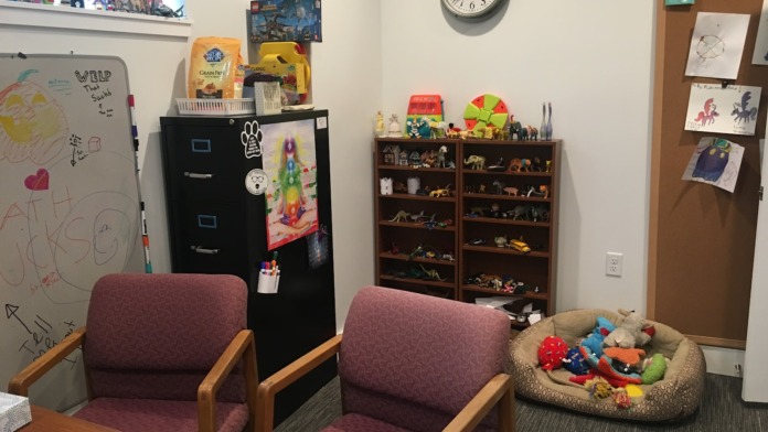 Interior playroom with purple chairs, white shelving units, toys, crafts, wall decorations, file storage, and whiteboard.