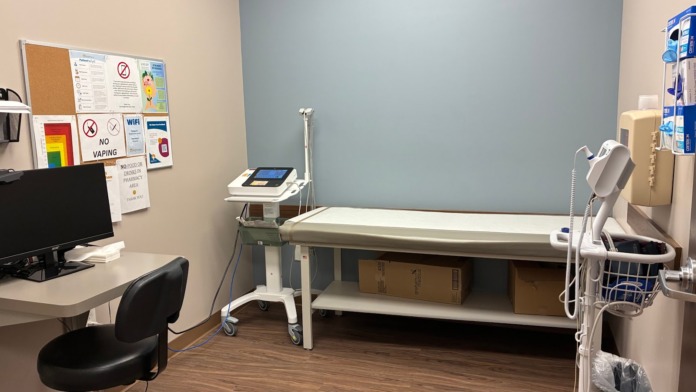 Interior clinical space with exam table, overhead light, computer workstation, equipment storage, and wall notices