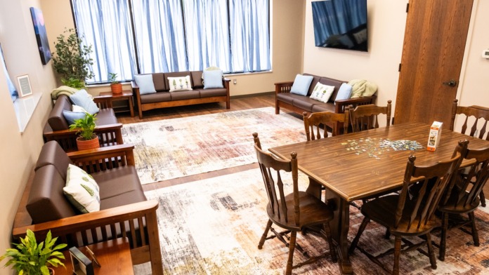 Interior multi-purpose room with dining table, wooden chairs, brown sofas, television, potted plants, and windows.