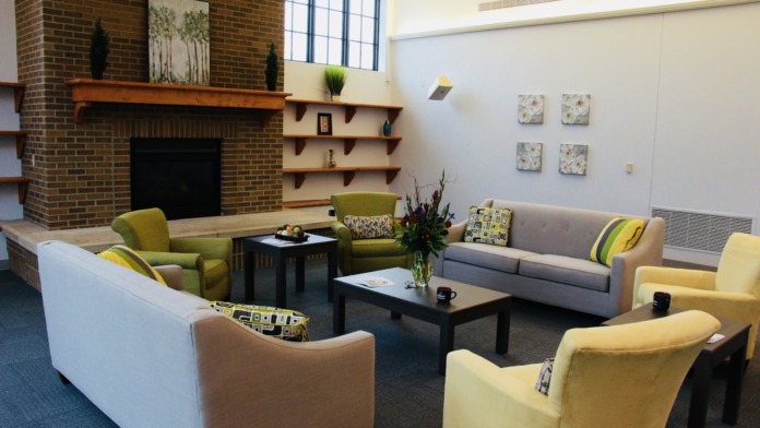 Interior lounge area with brick fireplace, wood mantel, floating shelves, colorful accent chairs, gray sofa, coffee table, and decorative artwork