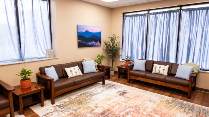 Interior lounge with brown leather sofas, wooden tables, area rug, landscape artwork, and large windows.