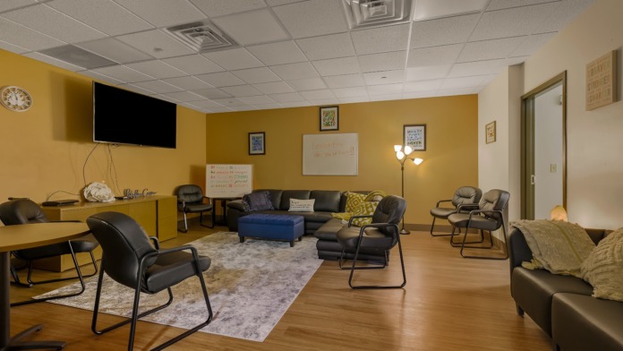 Interior community space with yellow walls, black leather seating, blue ottoman, mounted TV, whiteboards, and wooden flooring